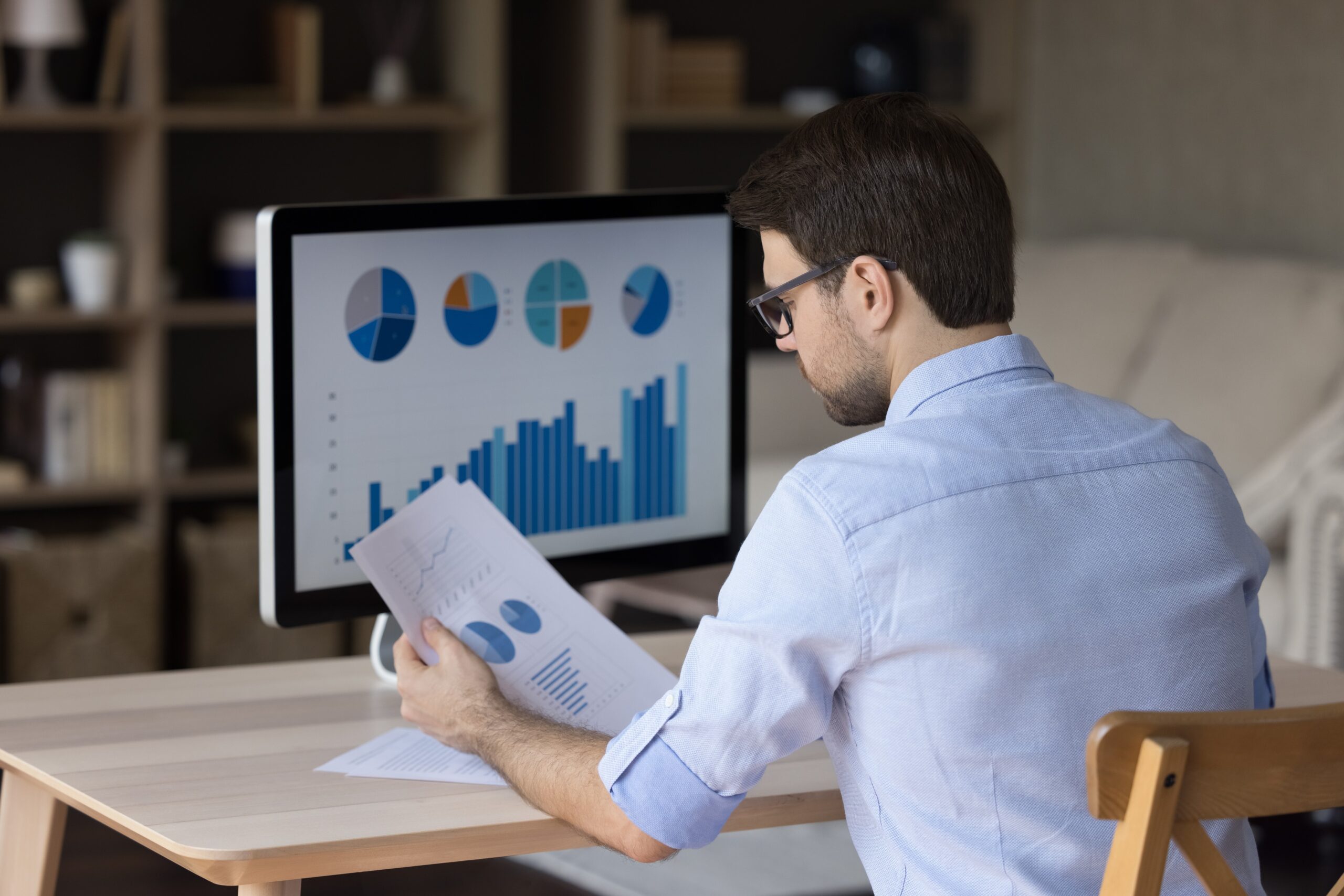 focused-millennial-business-man-analyzing-marketing-reports-desktop-monitor-reviewing-paper-graphs-financial-stats-startup-project-infographics-marketer-working-from-home-office-back-view